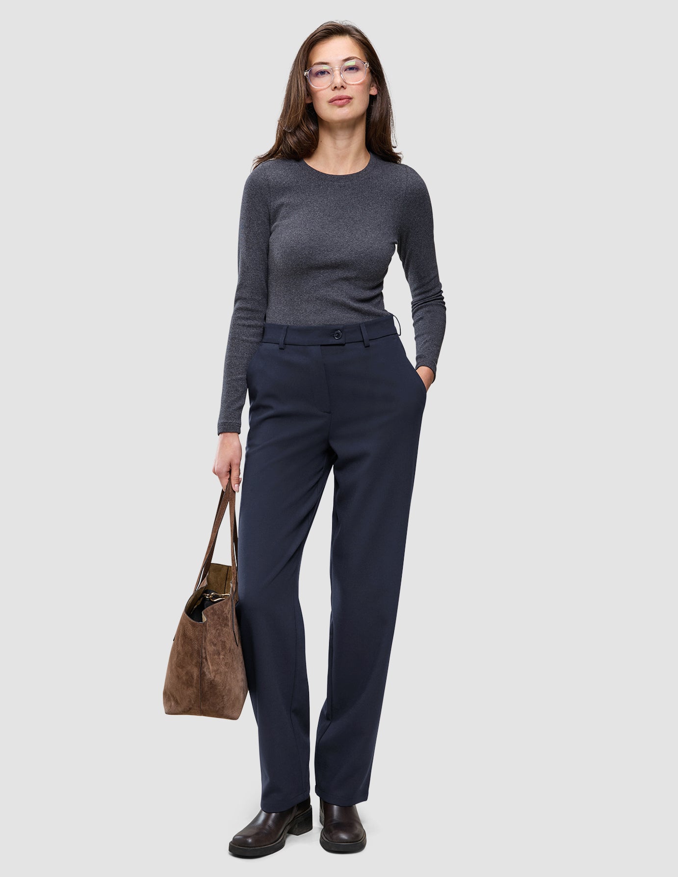 Eine Frau in einem dunkelgrauen Langarmhemd aus Doubleface-Stoff und einer Women GO2 Pants Straight Blue Nights, die eine braune Wildleder-Tragetasche bei sich trägt.